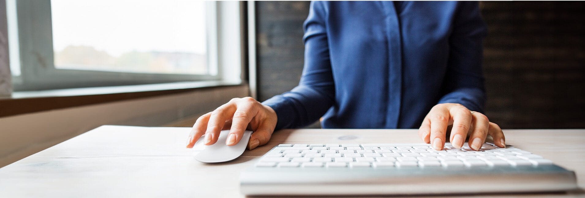 A woman is focused on typing on a keyboard while using a mouse with her right hand.