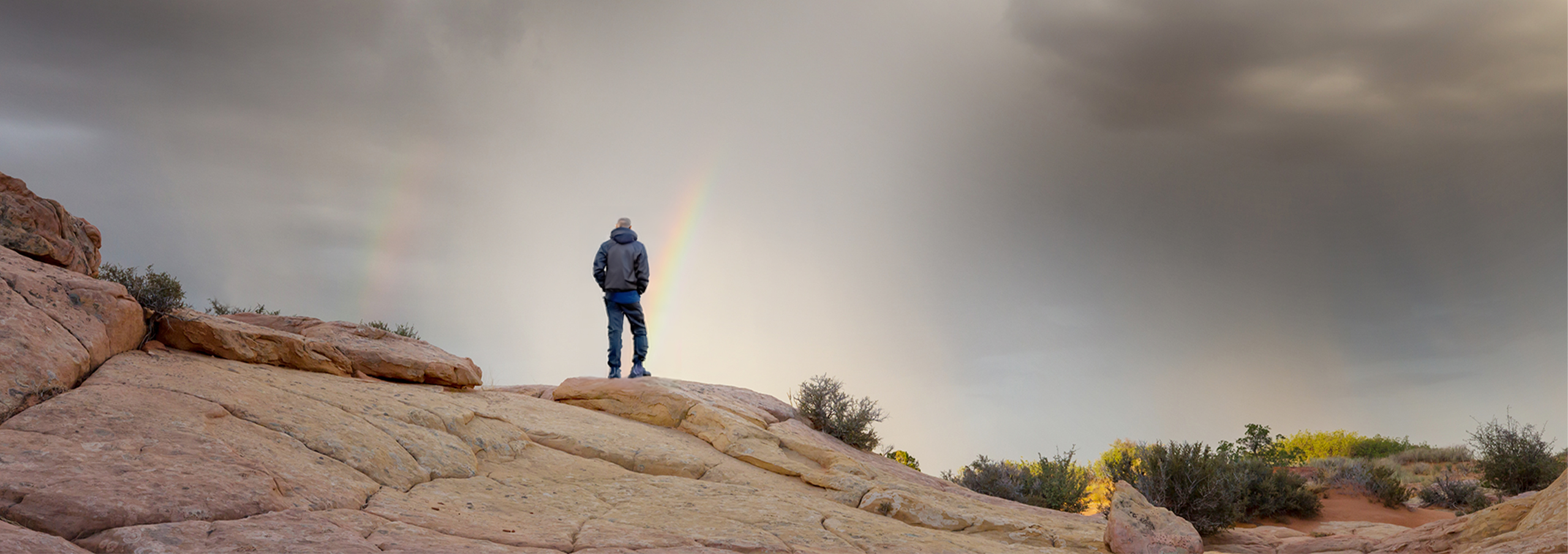 A person stands on a rock beneath a vibrant rainbow arching across a clear blue sky.
