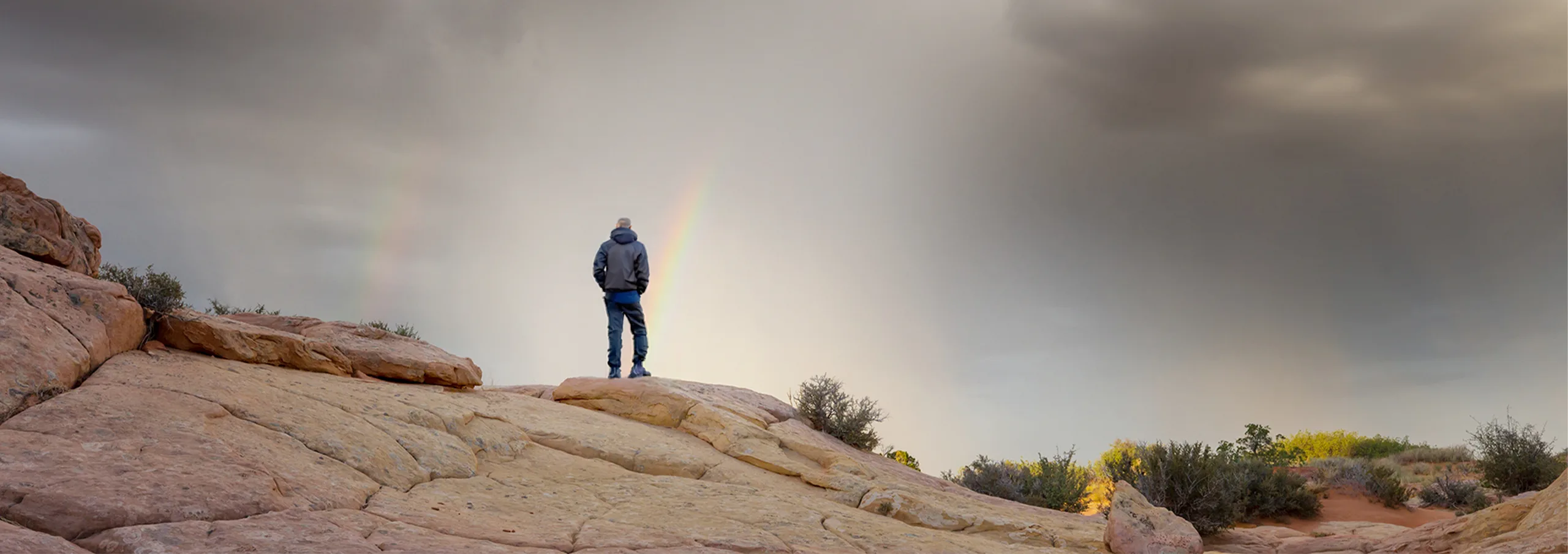 A person stands on a rock beneath a vibrant rainbow arching across a clear blue sky.